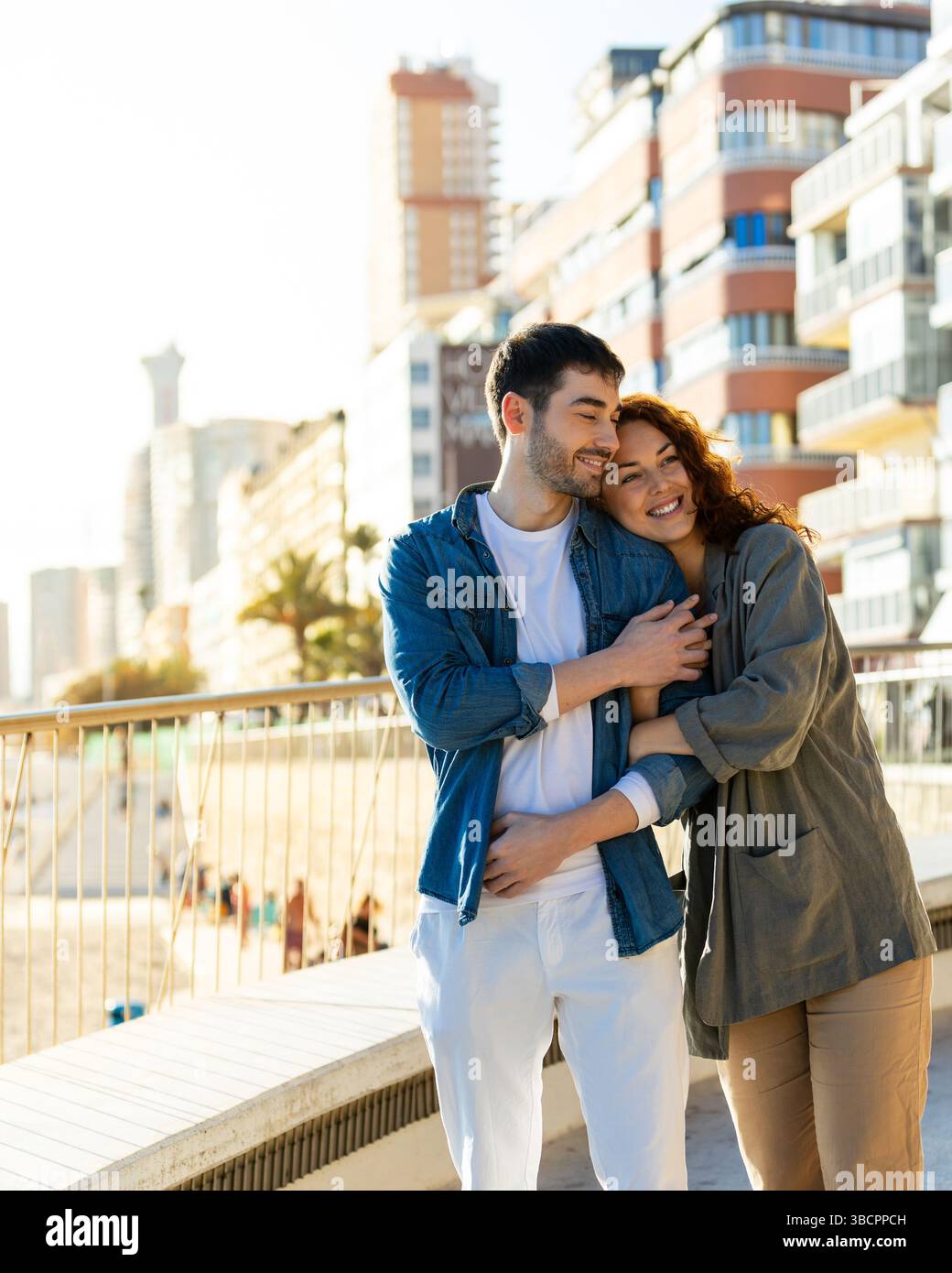 Young couple shares a cozy embrace on a beachfront walkway, smiling warmly against a backdrop of tall modern buildings and sunny coastal vibes. Lifest Stock Photo