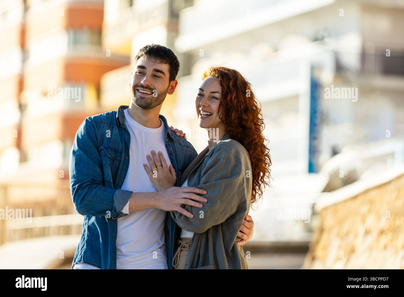 Joyful young couple enjoying a sunny moment on a city promenade, smiling and sharing a warm embrace in front of modern buildings and soft natural ligh Stock Photo