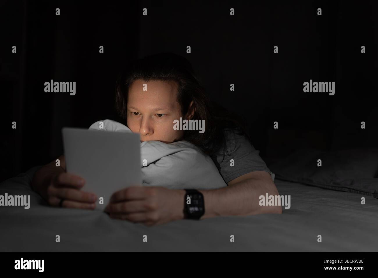 Man resting on bed, holding a tablet while browsing or reading in the dark with focused eyes. Stock Photo