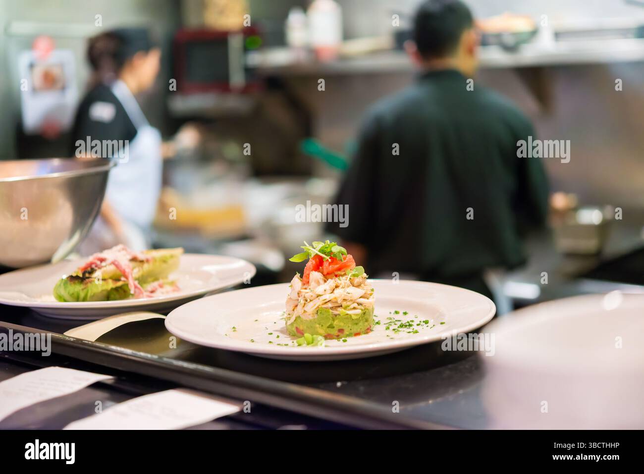 Restaurant Kitchen Food Expediting Counter // Plated dishes sit on the expediting counter between a busy restaurant kitchen and dining area, awaiting server pickup for delivery to guests. The finished plates, arranged in sequence of table orders, represent the critical transition point between back-of-house preparation and front-of-house service. This pass area, often supervised by an expediter who ensures quality control and proper timing, serves as the final checkpoint before meals reach diners in the restaurant. Stock Photo