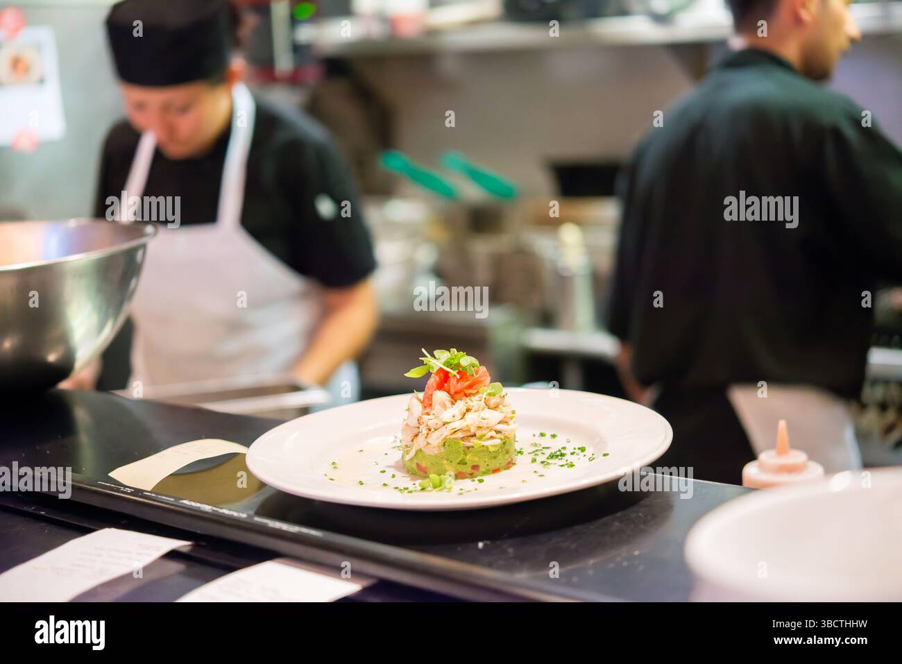 Restaurant Kitchen Food Expediting Counter Plated Dish // Plated dishes sit on the expediting counter between a busy restaurant kitchen and dining area, awaiting server pickup for delivery to guests. The finished plates, arranged in sequence of table orders, represent the critical transition point between back-of-house preparation and front-of-house service. This pass area, often supervised by an expediter who ensures quality control and proper timing, serves as the final checkpoint before meals reach diners in the restaurant. Stock Photo