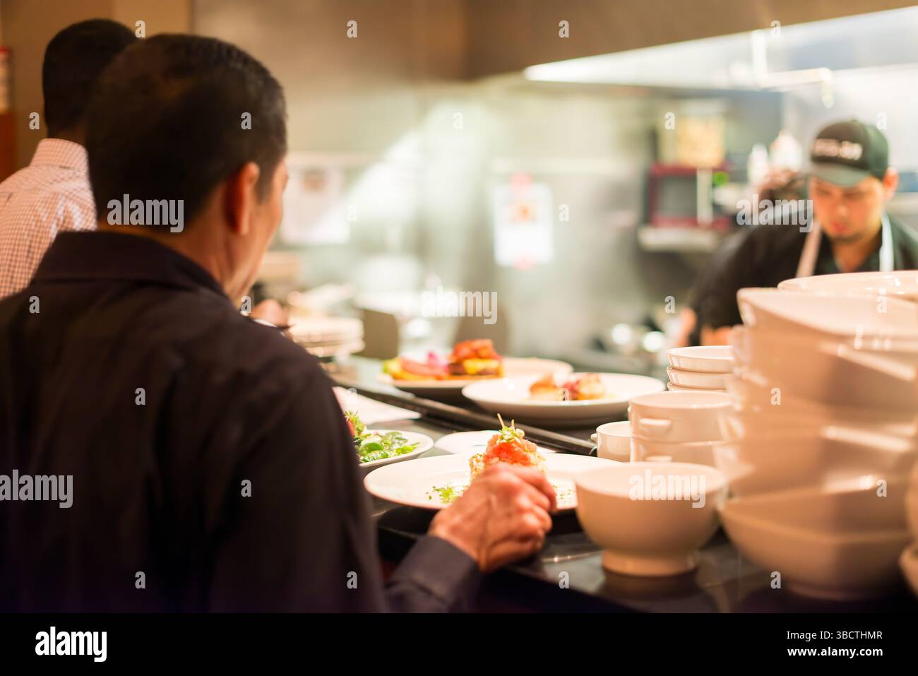 Restaurant Kitchen Expediting Counter Plated Dishes // Plated dishes sit on the expediting counter between a busy restaurant kitchen and dining area, awaiting server pickup for delivery to guests. The finished plates, arranged in sequence of table orders, represent the critical transition point between back-of-house preparation and front-of-house service. This pass area, often supervised by an expediter who ensures quality control and proper timing, serves as the final checkpoint before meals reach diners in the restaurant. Stock Photo