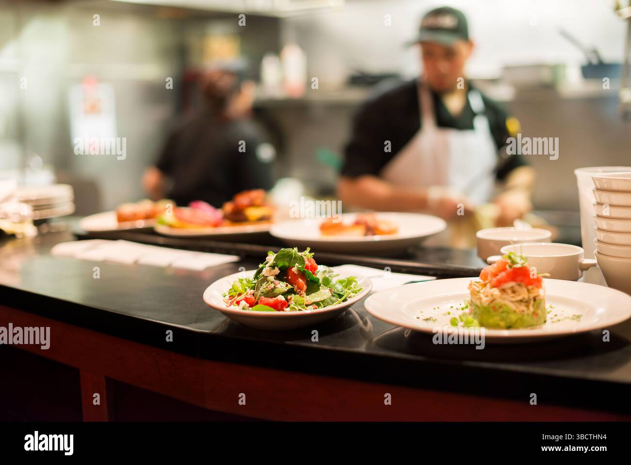 Restaurant Kitchen Food Expediting Counter // Plated dishes sit on the expediting counter between a busy restaurant kitchen and dining area, awaiting server pickup for delivery to guests. The finished plates, arranged in sequence of table orders, represent the critical transition point between back-of-house preparation and front-of-house service. This pass area, often supervised by an expediter who ensures quality control and proper timing, serves as the final checkpoint before meals reach diners in the restaurant. Stock Photo