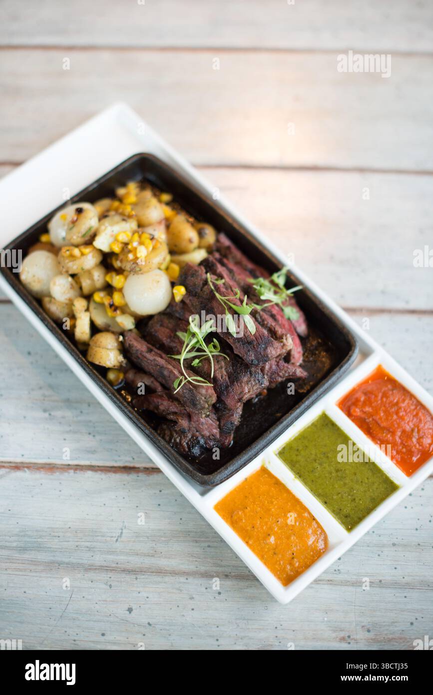 Seared Steak With Potatoes And Salsa Sauces // Strips of seared steak arranged on a plate accompanied by baby potatoes and corn kernels. The Mexican-inspired dish features a trio of colorful sauces: a vibrant orange salsa, a fresh green sauce reminiscent of salsa verde, and a deep red salsa. The presentation offers a deconstructed take on traditional Mexican flavors in an elegant, plated format. Stock Photo