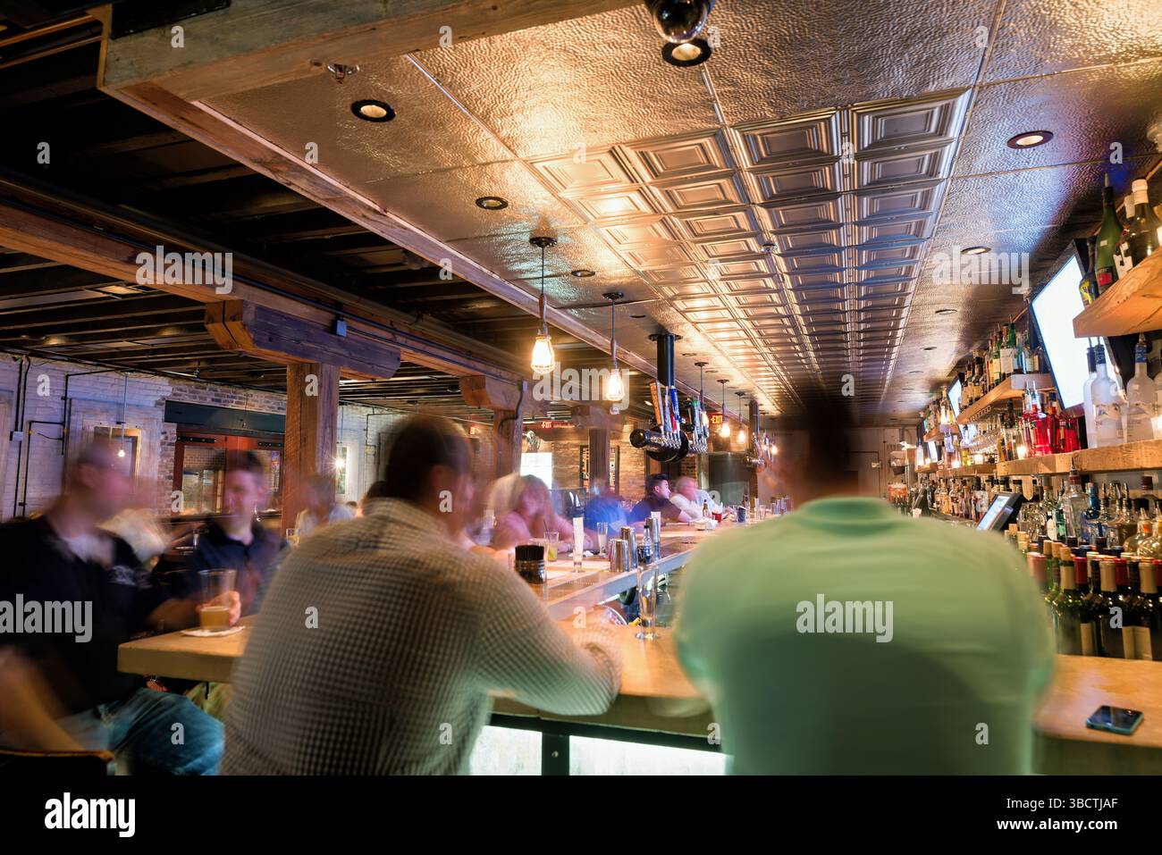 Virtue Feed And Grain Bar Alexandria Virginia // ALEXANDRIA, Virginia — The busy bar at Virtue Feed and Grain restaurant buzzes with activity in Old Town Alexandria. Located in a renovated 19th-century warehouse that once served as a feed and grain store, the bar area features industrial-inspired design elements that honor the building's utilitarian past. Patrons gather at the wooden bar counter where bartenders prepare craft cocktails and serve beverages in this popular Old Town establishment. Stock Photo