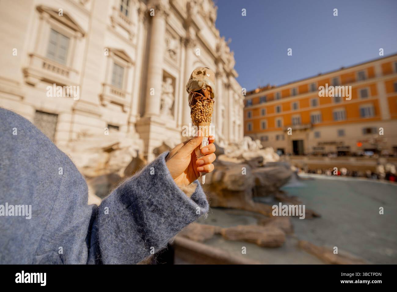 Close-up of a hand holding a gelato cone with Trevi Fountain softly blurred in the background, capturing a sweet Roman moment in golden light Stock Photo