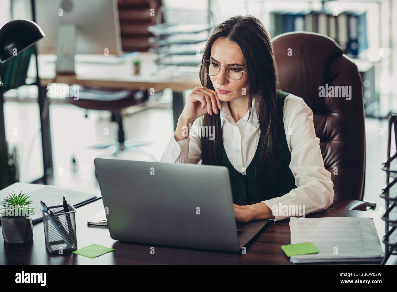 Experienced professional businesswoman focused on her work, analyzing documents and using a laptop in modern office interior Stock Photo