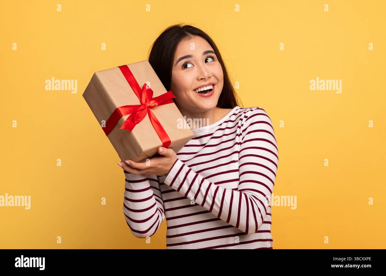 Joyful woman holding a gift with a red ribbon against a bright yellow background Stock Photo