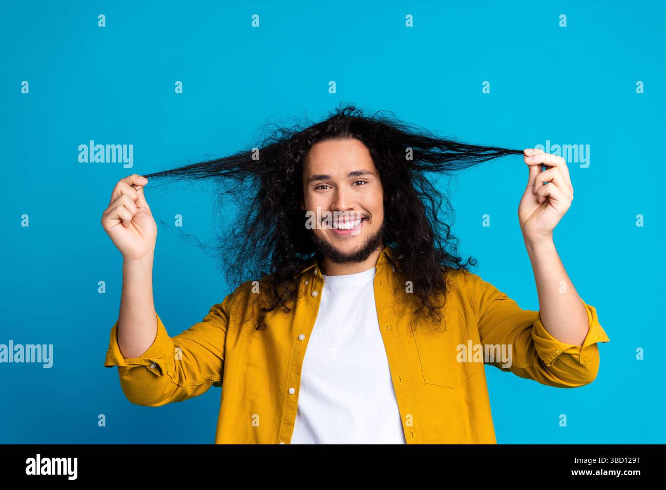 Cheerful man with curly hair in yellow shirt smiling against vibrant blue studio background Stock Photo