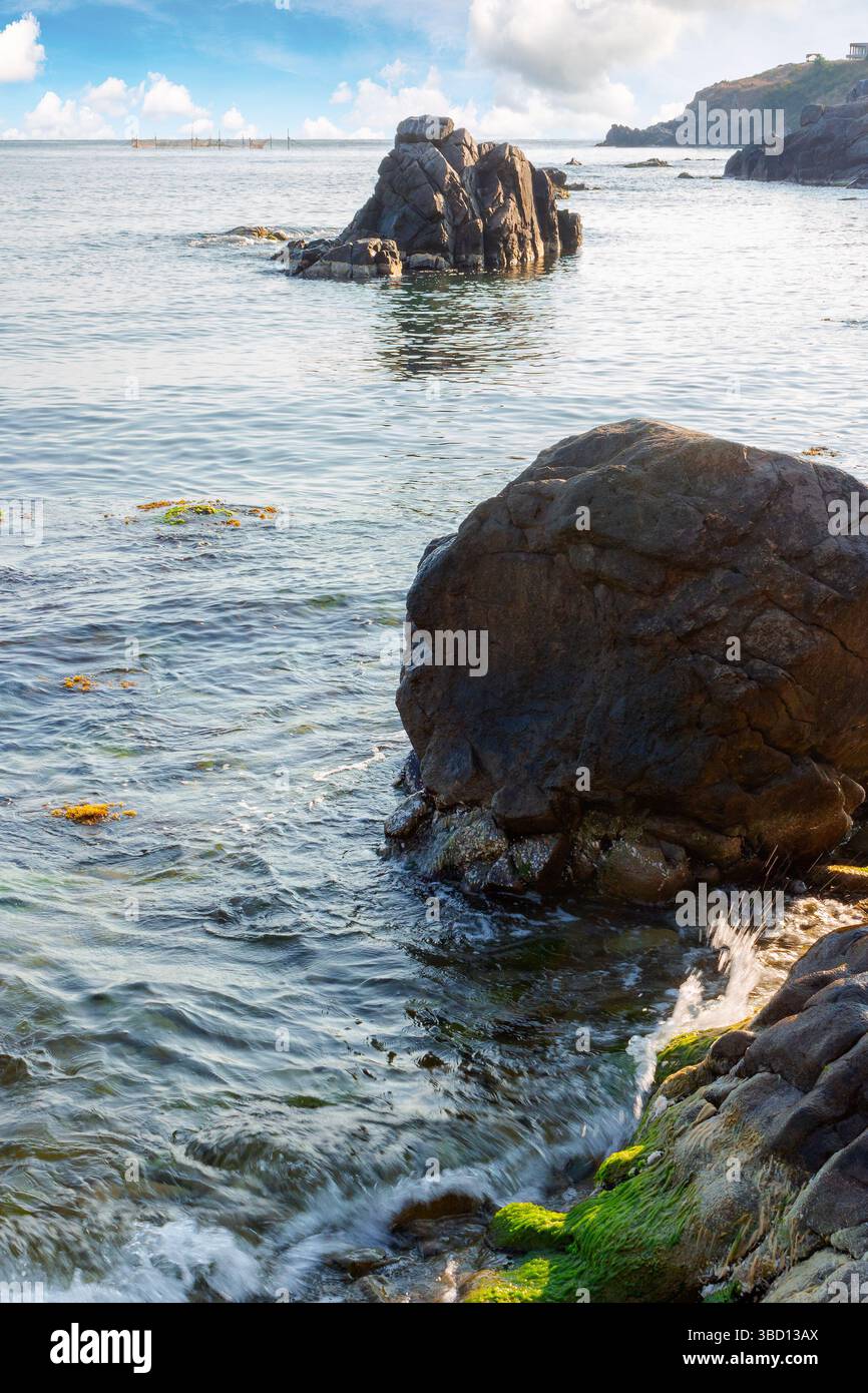 seascape with rock in the water. textured boulder reflecting calm sea surface in morning light. vacation season in summer. sunny weather. fluffy cloud Stock Photo