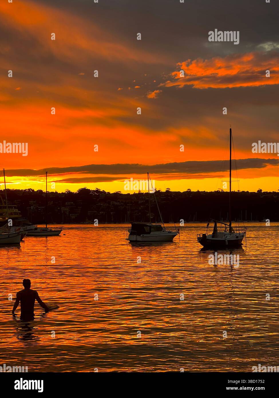 Striking sunset over a calm bay with silhouetted boats anchored in still water and a lone person wading in the glowing orange reflection - Smartphone Captured Stock Image