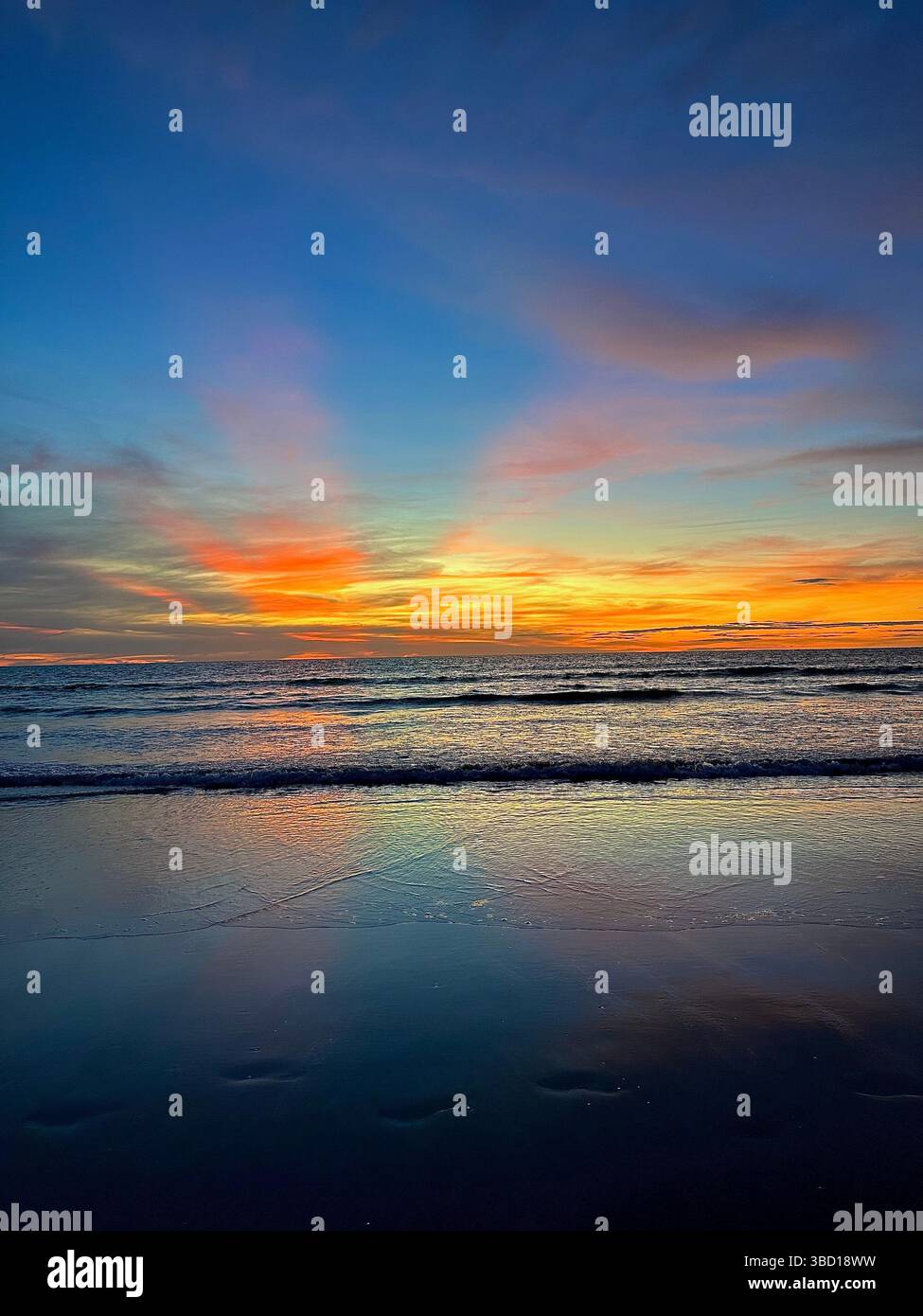 Vibrant ocean sunset with radiant hues of orange, pink, and blue reflecting on calm waves and wet sand, creating a tranquil and inspiring scene - Smartphone Captured Stock Image