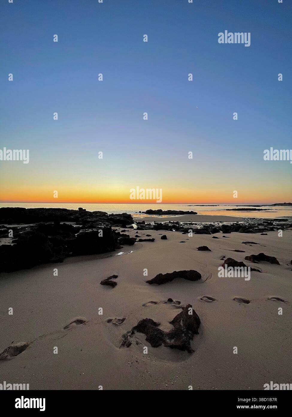 Tranquil beach at sunrise with footprints in the sand and rocky shoreline—perfect for travel, wellness, solitude, and coastal lifestyle themes - Smartphone Captured Stock Image