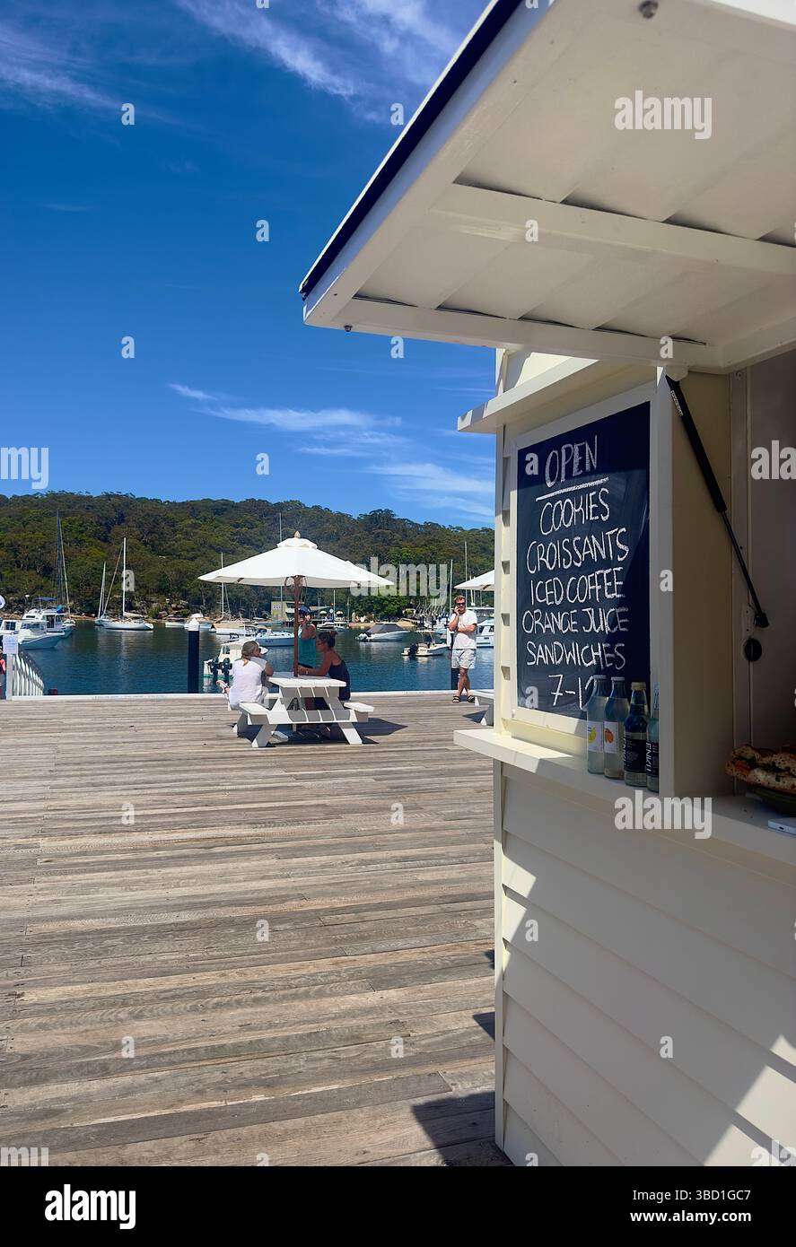 Seaside café kiosk on a sunny marina boardwalk with outdoor seating, sailboats in the harbor, and people enjoying the waterfront view. - Smartphone Captured Stock Image