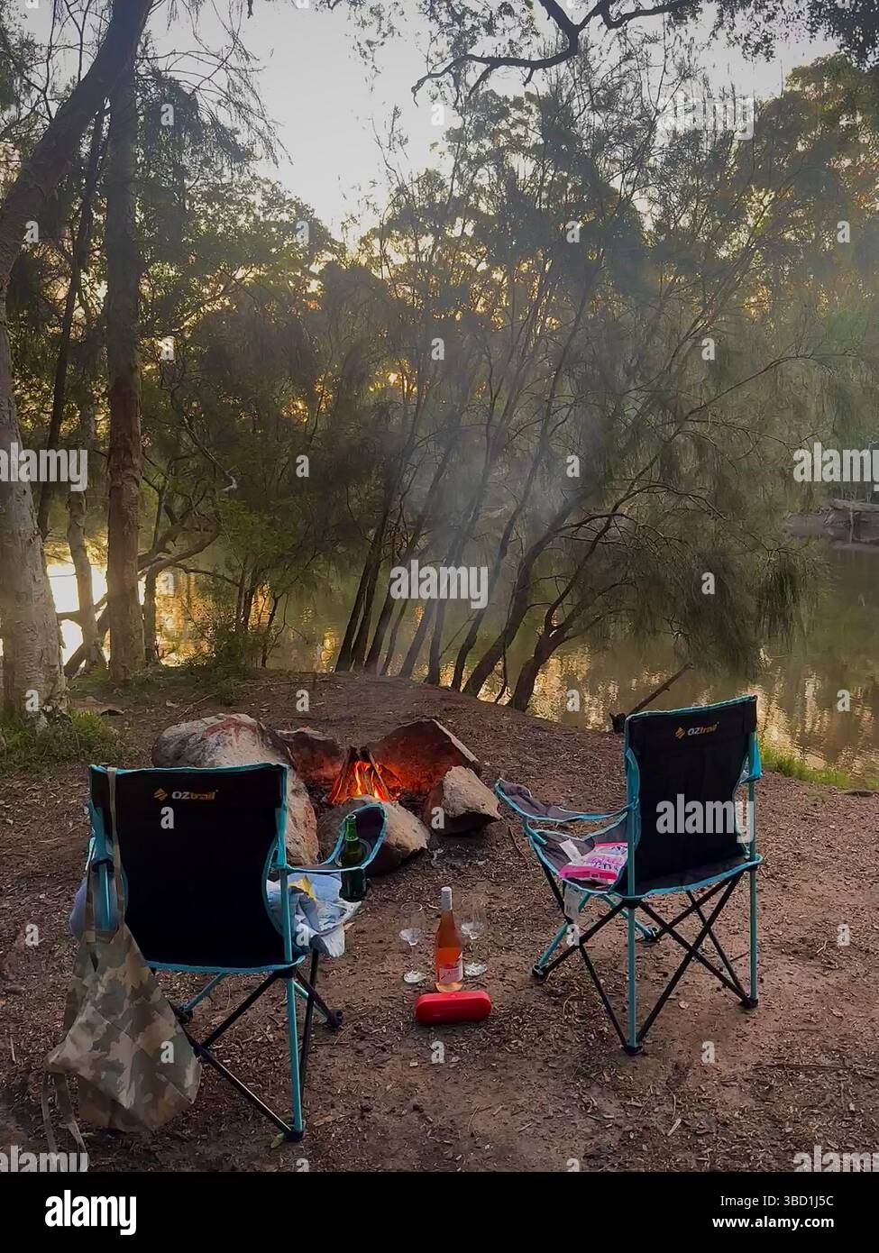 Cozy riverside campsite with two chairs around a campfire, overlooking calm water through the trees at sunset. - Smartphone Captured Stock Image