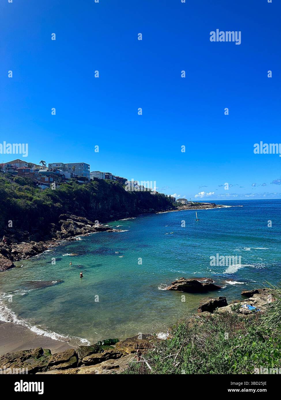 Turquoise bay nestled between rocky headlands under a vibrant blue sky, with calm sea waters and coastal homes overlooking the tranquil shoreline. - Smartphone Captured Stock Image