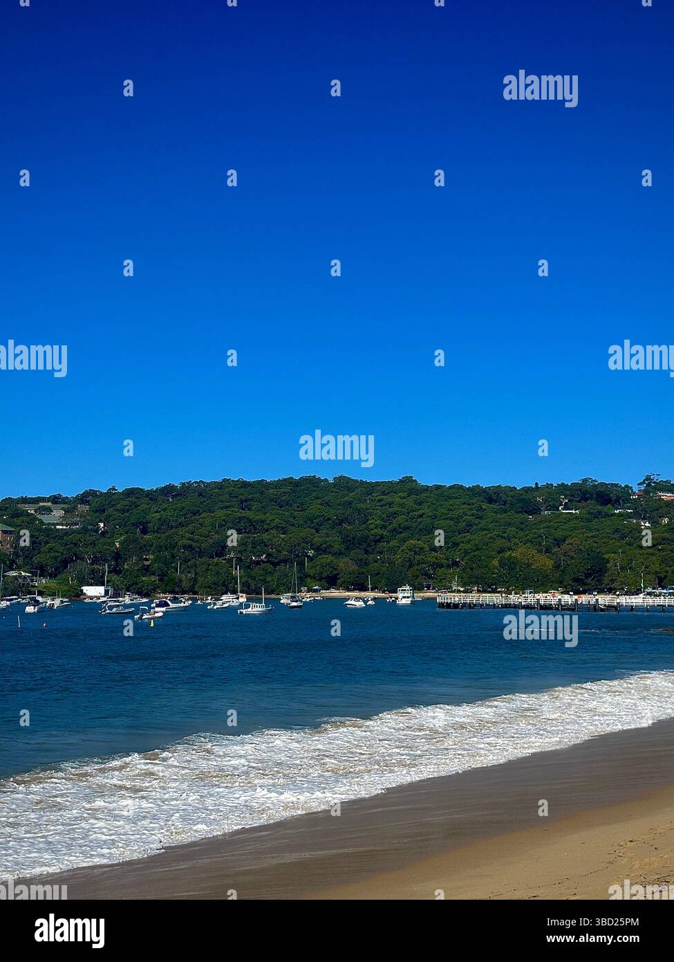 Scenic coastal view of sandy beach, gentle waves, and anchored boats under a clear blue sky in a tranquil seaside harbor - Smartphone Captured Stock Image