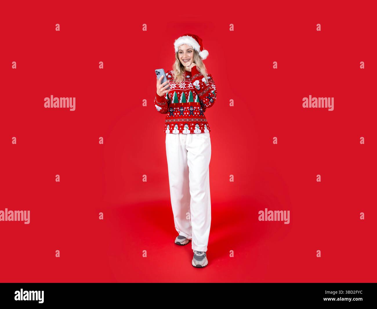 charming caucasian Santa woman holding phone. Smiling blonde girl looking, chatting on smartphone standing over isolated red background. Christmas. Stock Photo