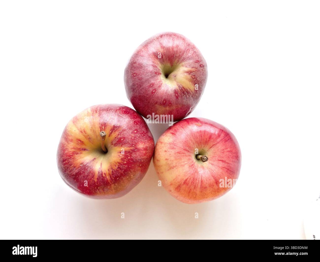 Three apples are sitting on a white surface. The apples are red and shiny. They are arranged in a triangular shape Stock Photo