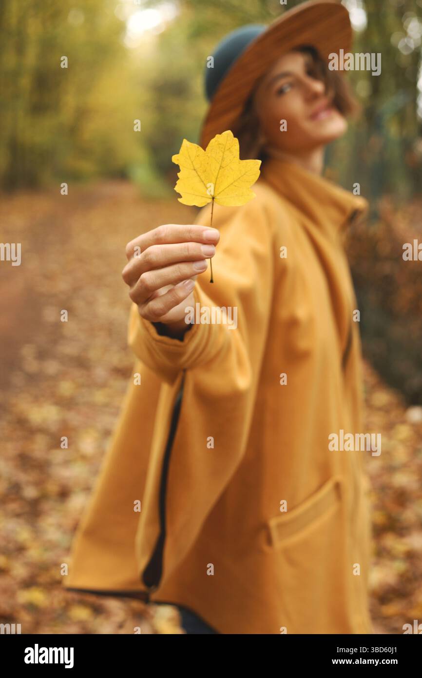 Beautiful woman holding autumn coloured leaf towards camera, while standing in scenic fall nature environment. Model not on focus, wearing hat, poncho Stock Photo