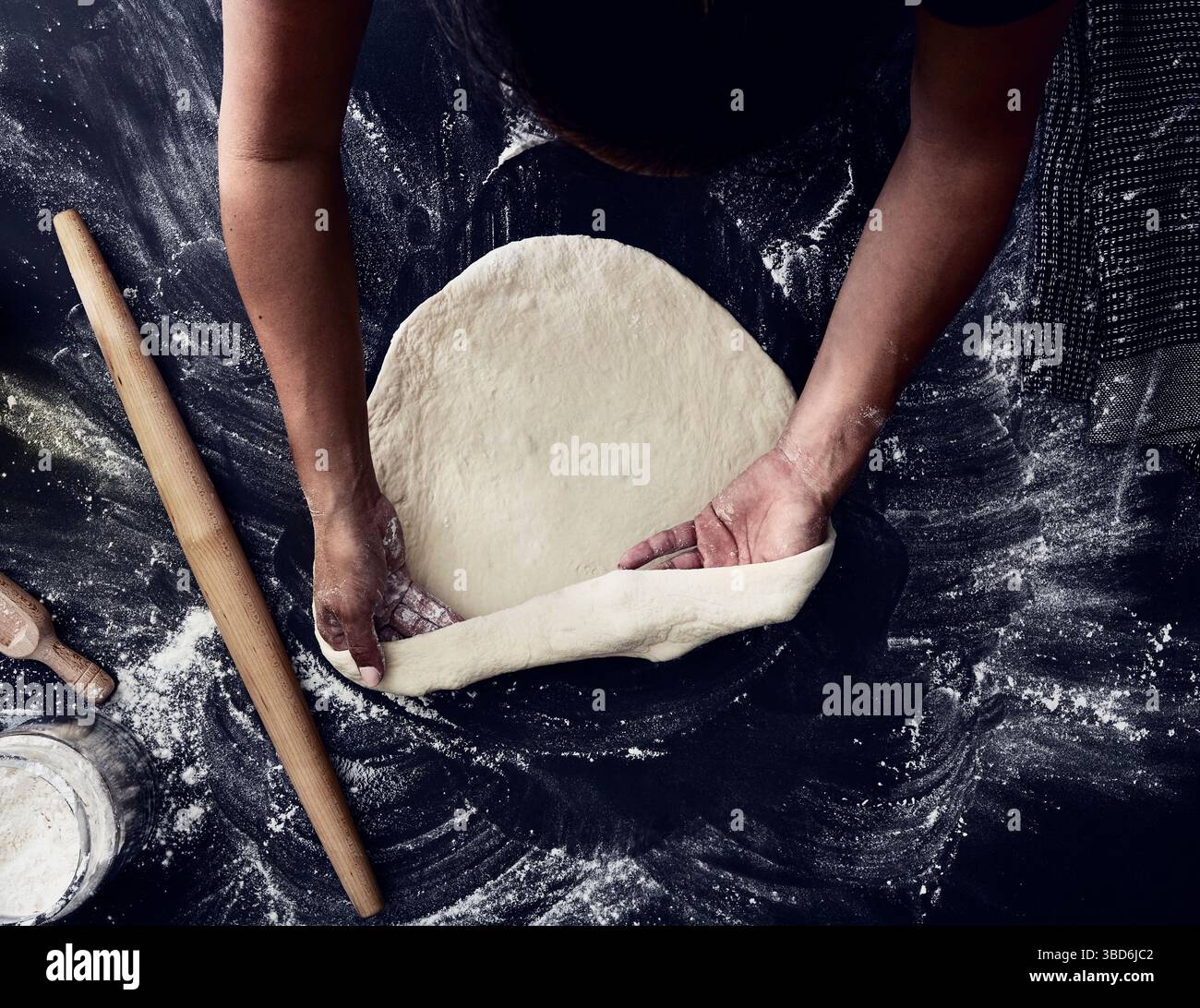 Person stretches dough on a dark surface dusted with flour. Stock Photo