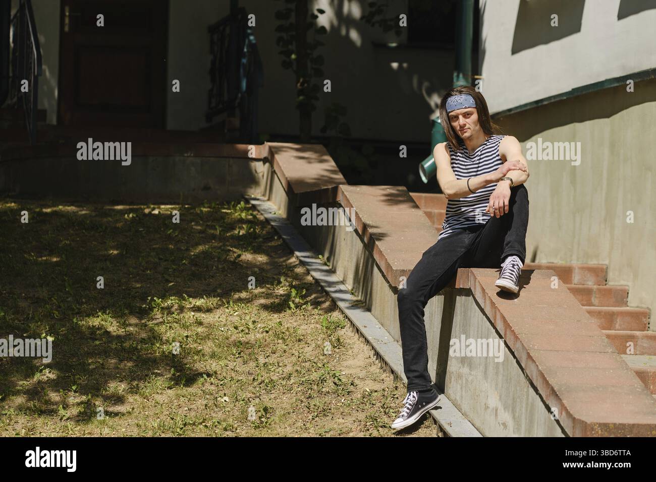 Creative long-haired man in tank top and jeans sits on railing Stock Photo