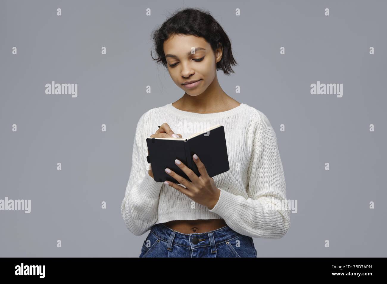 Concentrated young african woman standing against grey background and writing in diary Stock Photo