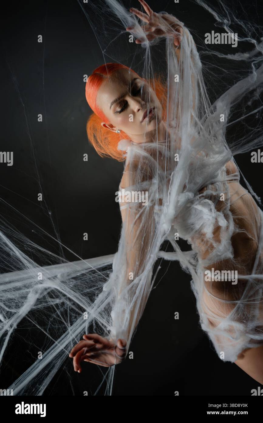 Red-haired woman entangled in an intricate net trap against a dark backdrop Stock Photo