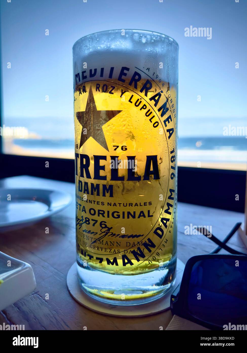 Tall glass of estrella beer with condensation on a table with a beach and ocean view in the background, - Smartphone Captured Stock Image