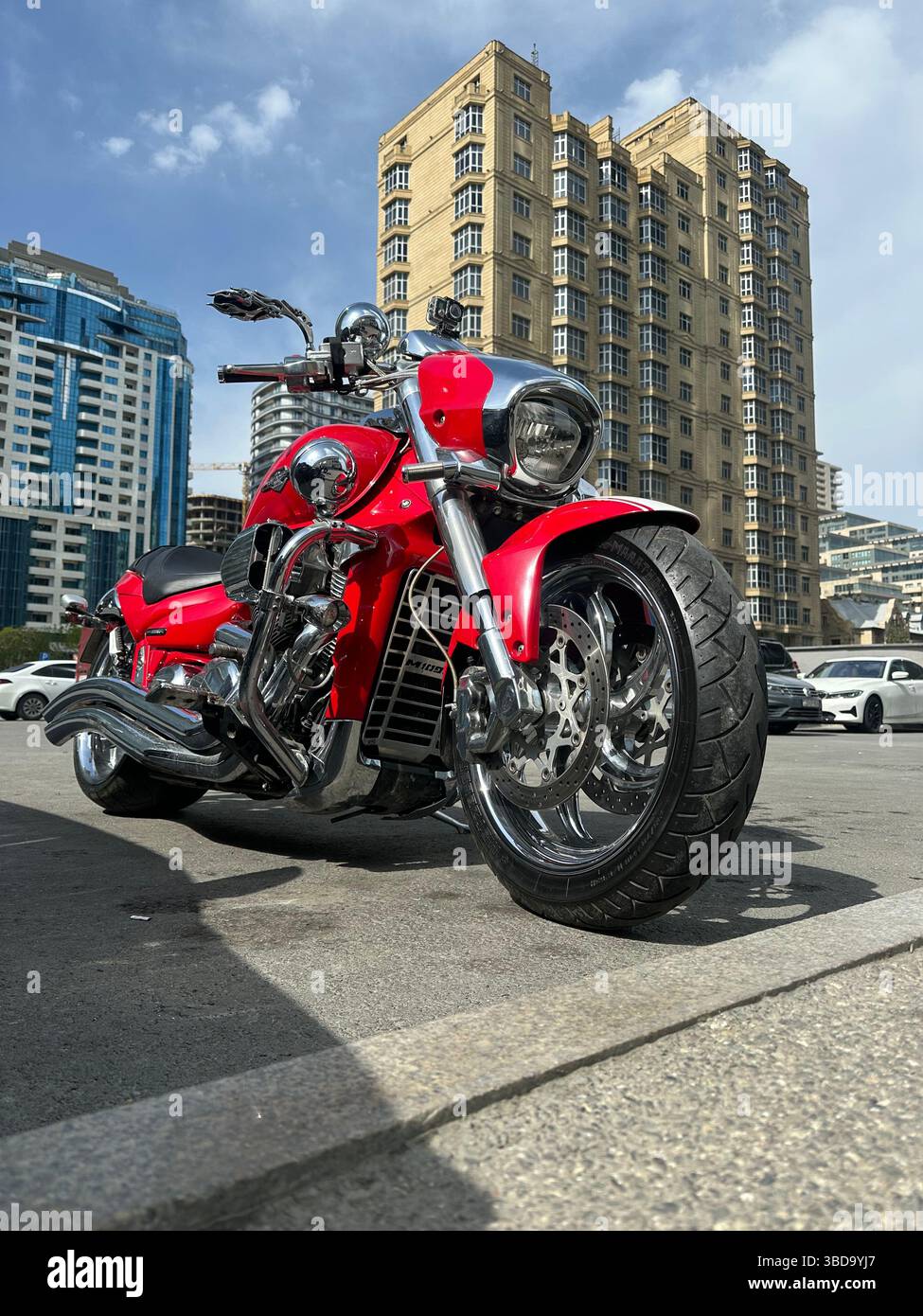 A striking red cruiser motorcycle parked in a modern cityscape, shining under the daylight with tall buildings and clear skies in the background - Smartphone Captured Stock Image