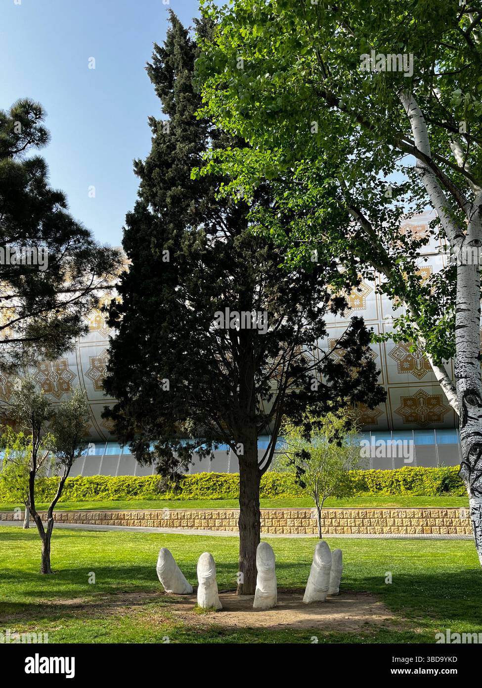 A unique park sculpture shows giant stone fingers emerging from the ground beneath a tall tree, blending art with nature in a peaceful green setting - Smartphone Captured Stock Image