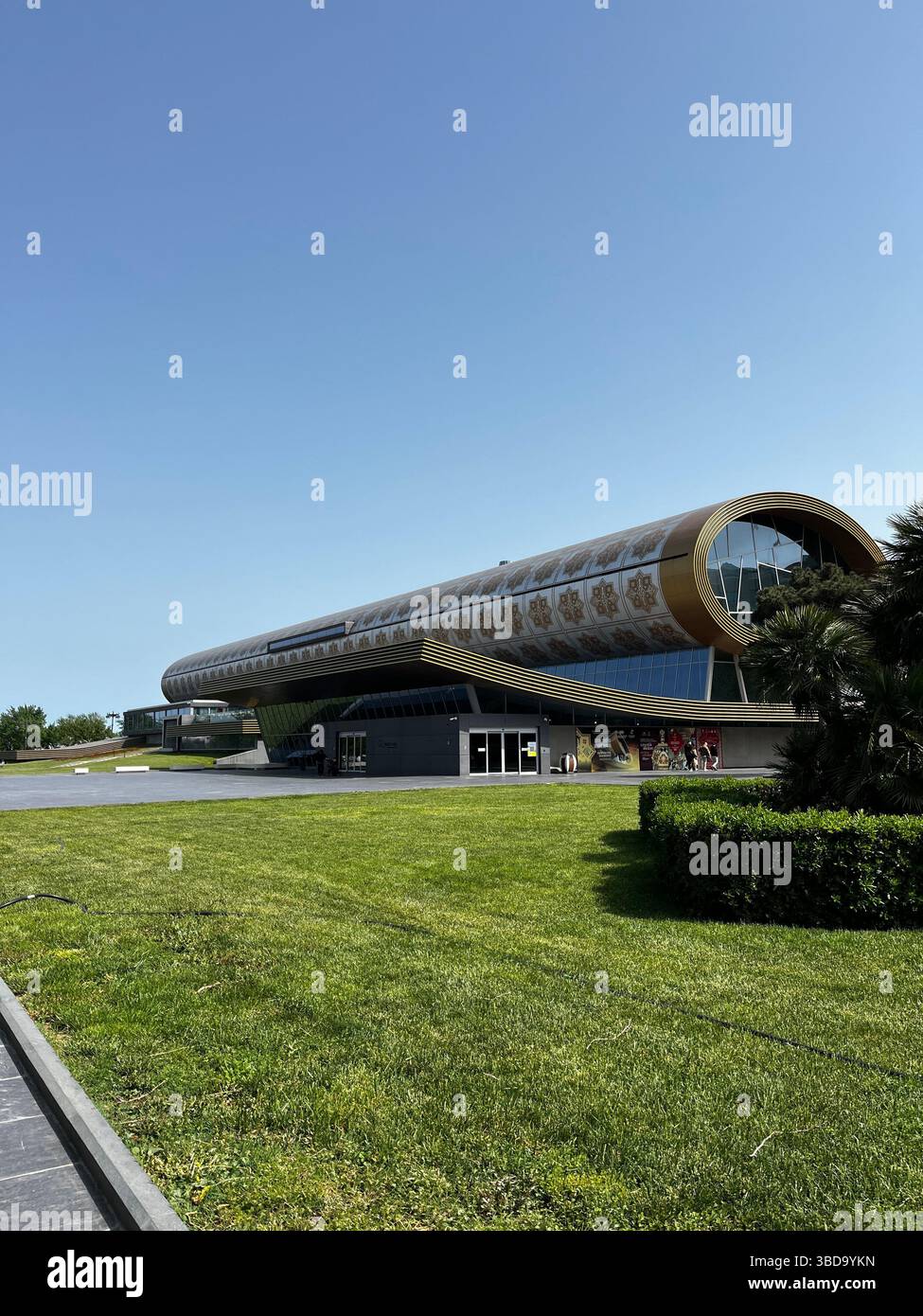 Modern cylindrical building with artistic exterior design under a clear blue sky, surrounded by a well-maintained lawn and palm trees, likely a museum - Smartphone Captured Stock Image