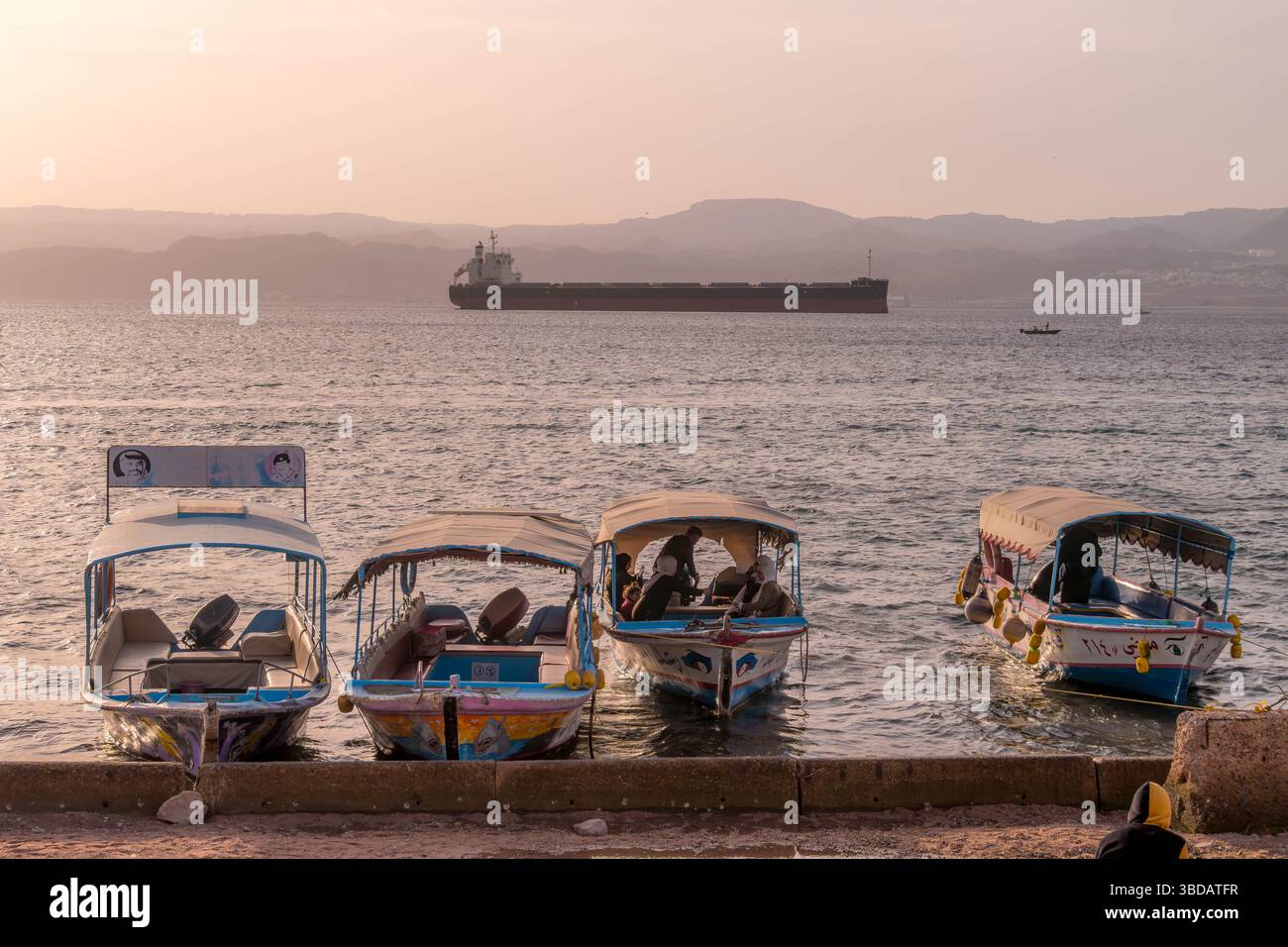 Tourist boats docked at sunset on the Aqaba shore, Persian Gulf, with a cargo ship and Israel mountains silhouettes in the Middle East panorama. Stock Photo