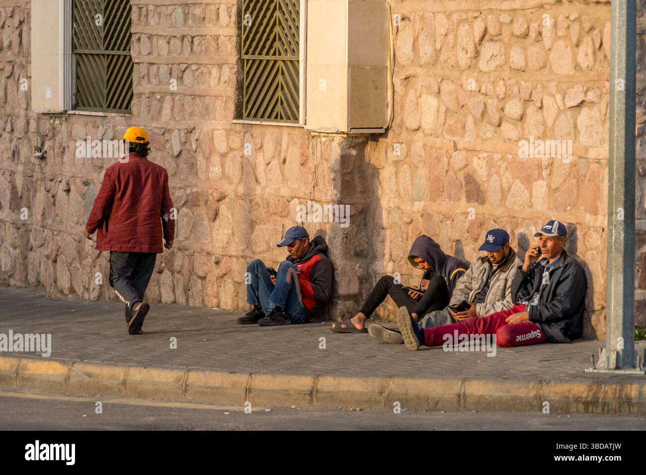 Several Arab Jordanian men sit idly on a city sidewalk in Aqaba, highlighting themes of unemployment, and social challenges. Stock Photo