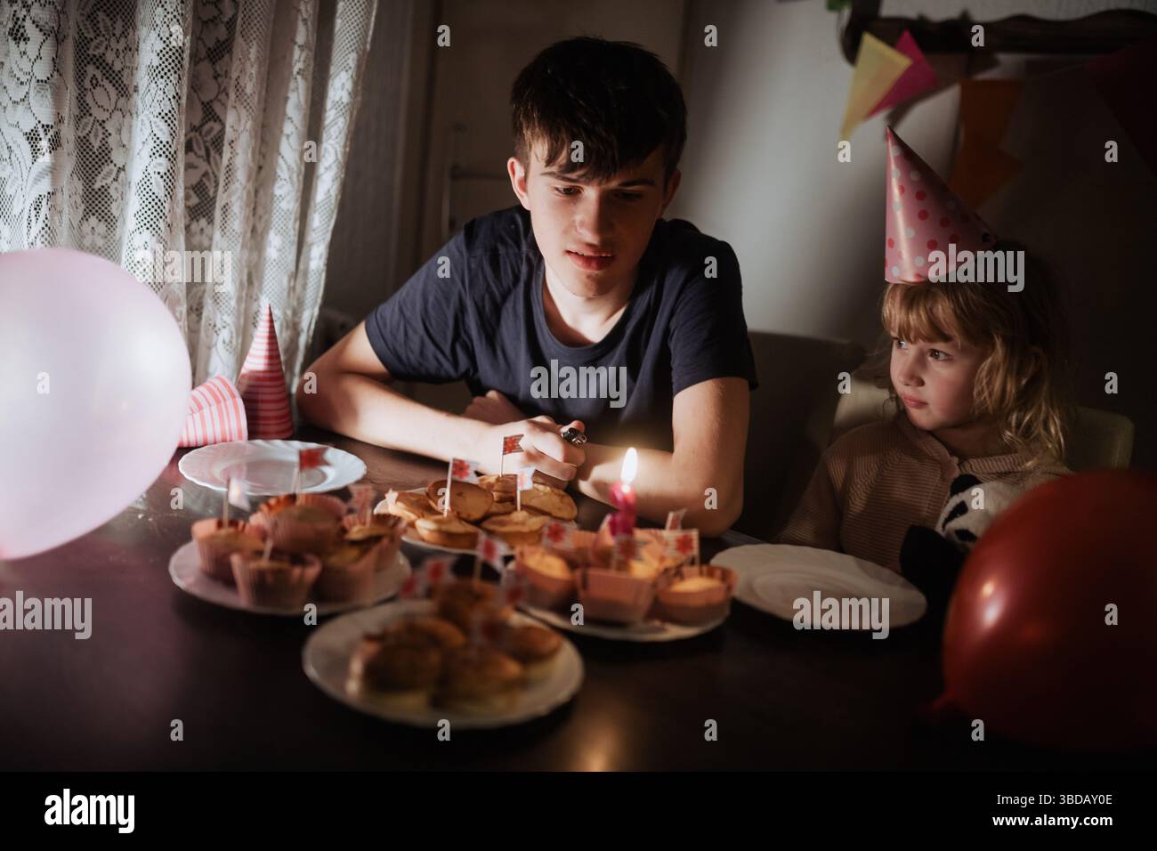 Two siblings enjoy a birthday celebration with a spread of colorful cupcakes and balloons. The atmosphere is festive, featuring party hats and soft li Stock Photo