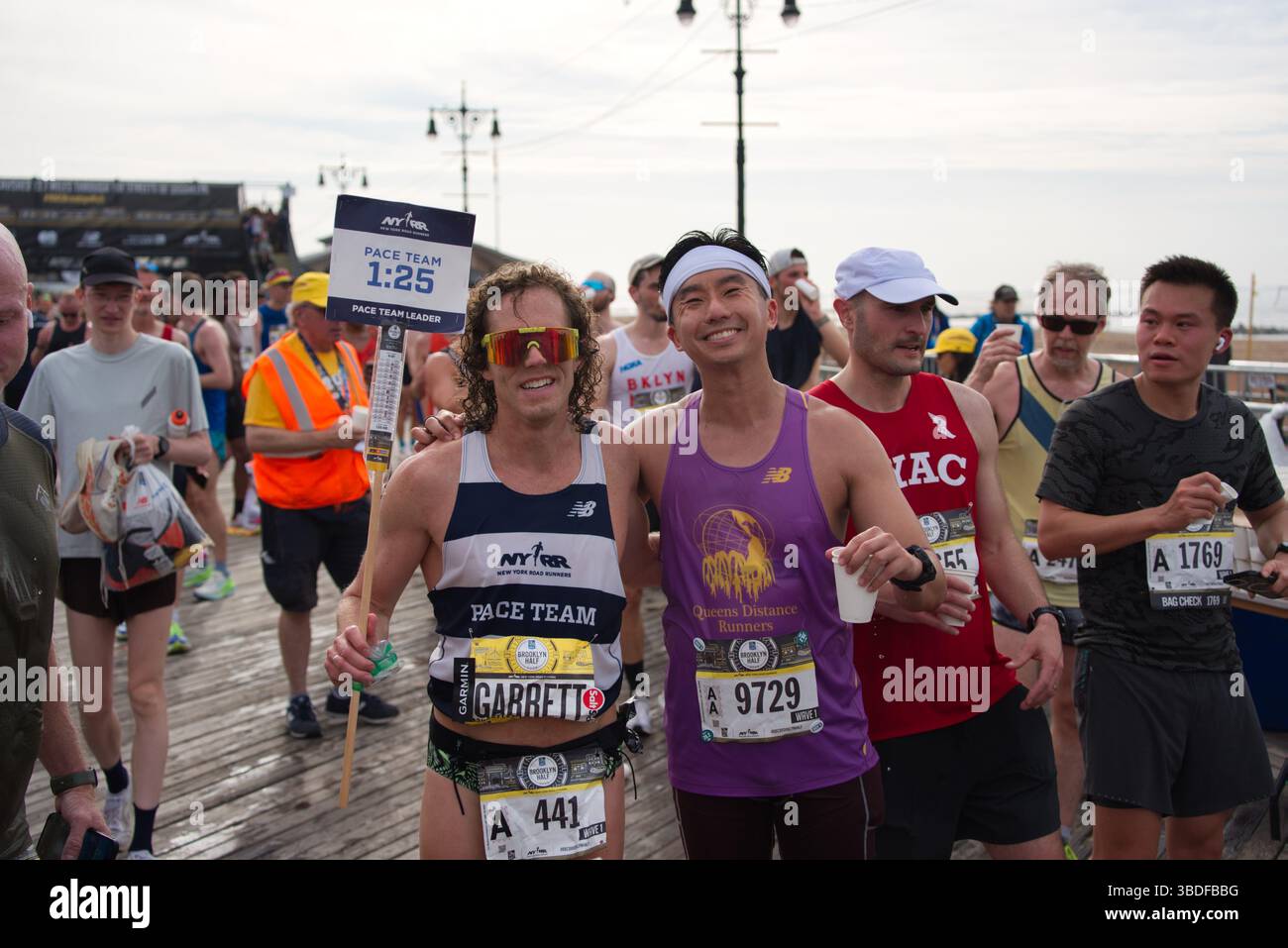 Two male runners, one wearing a navy blue and white singlet and the other a purple singlet, stand together at a marathon.  They are smiling and holdin Stock Photo