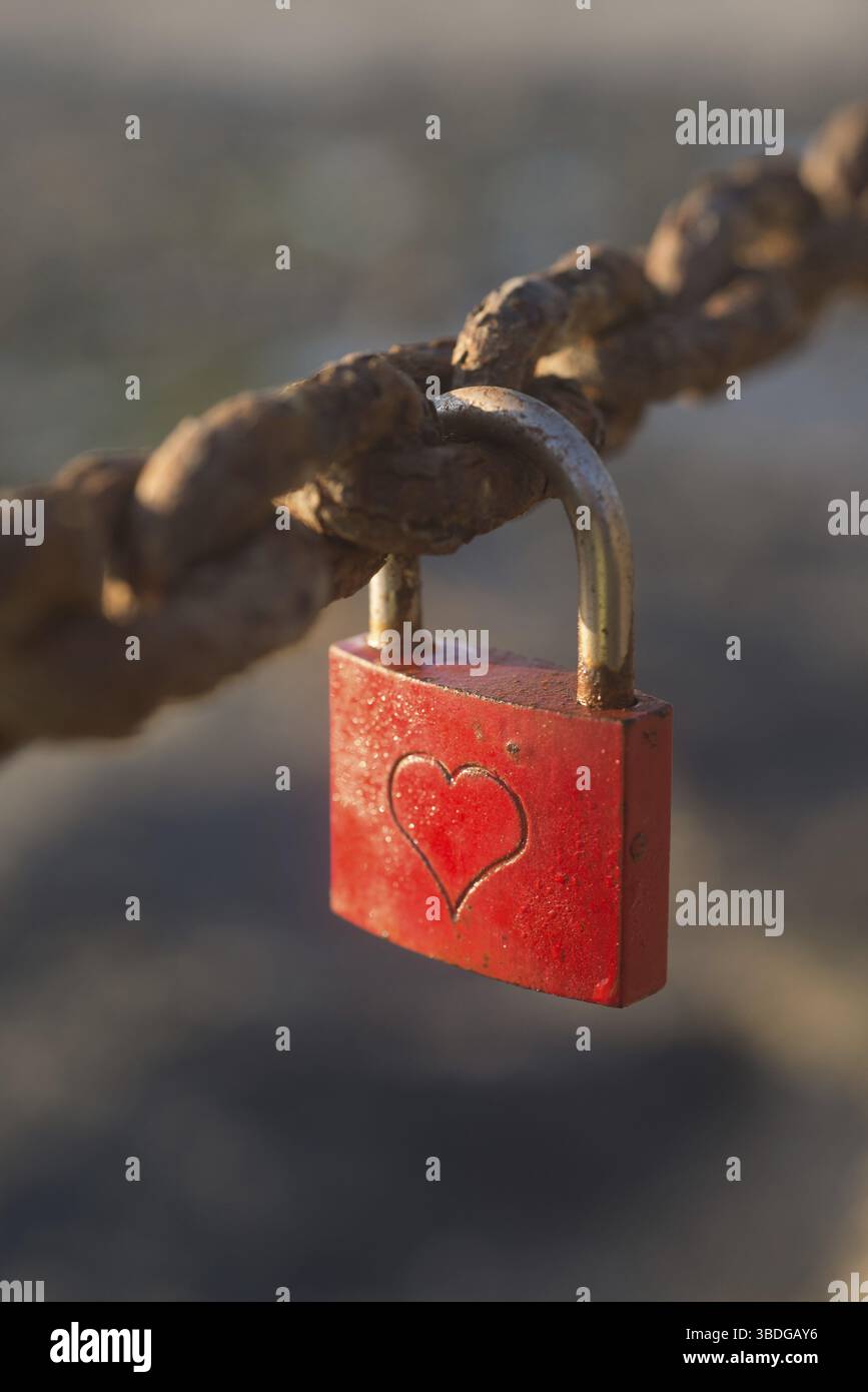 A small lock, painted red, with a heart engraved on it, is locked on to a rusty chain Stock Photo