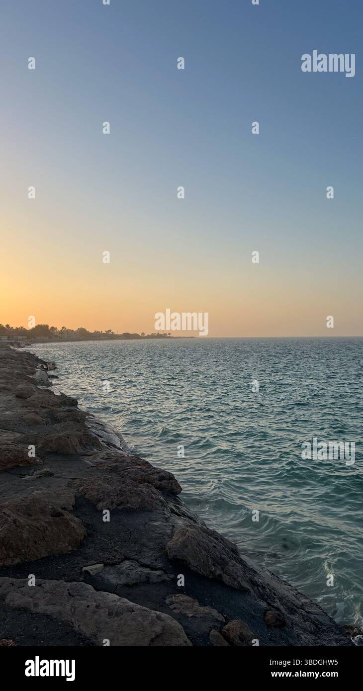A rocky shoreline meets calm, turquoise sea waters under a soft orange and blue sunset sky. The distant coastline is dotted with trees and buildings. - Smartphone Captured Stock Image