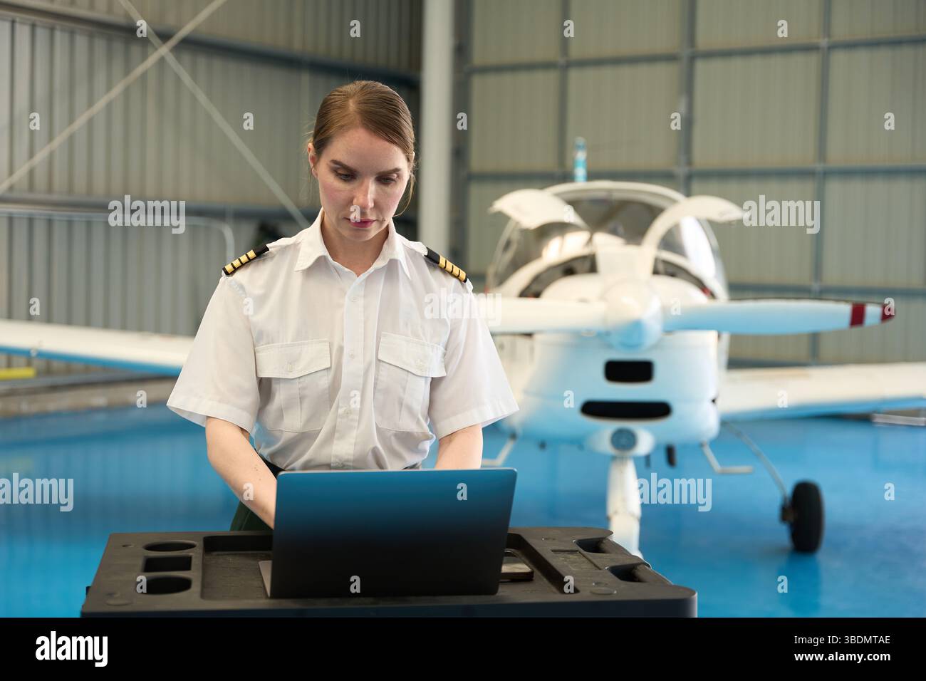 Female pilot working on laptop in aircraft hangar with small plane Stock Photo