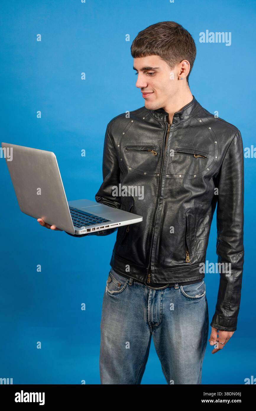 Young man with black leather jacket holding a laptop and smiling on a blue background Stock Photo
