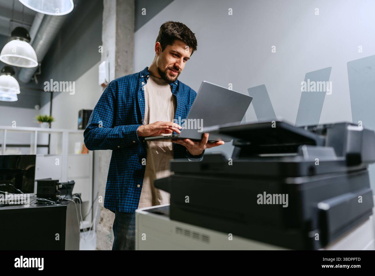 Man using laptop in modern office space while standing next to printer during working hours Stock Photo