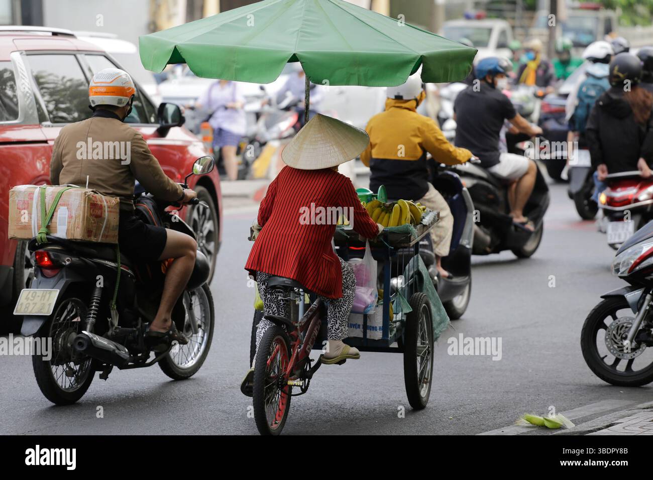 Ho Chi Minh, Vietnam - December 26, 2022: Vietnamese street fruit vendor rides his bike wearing a conical hat, Non La. Stock Photo