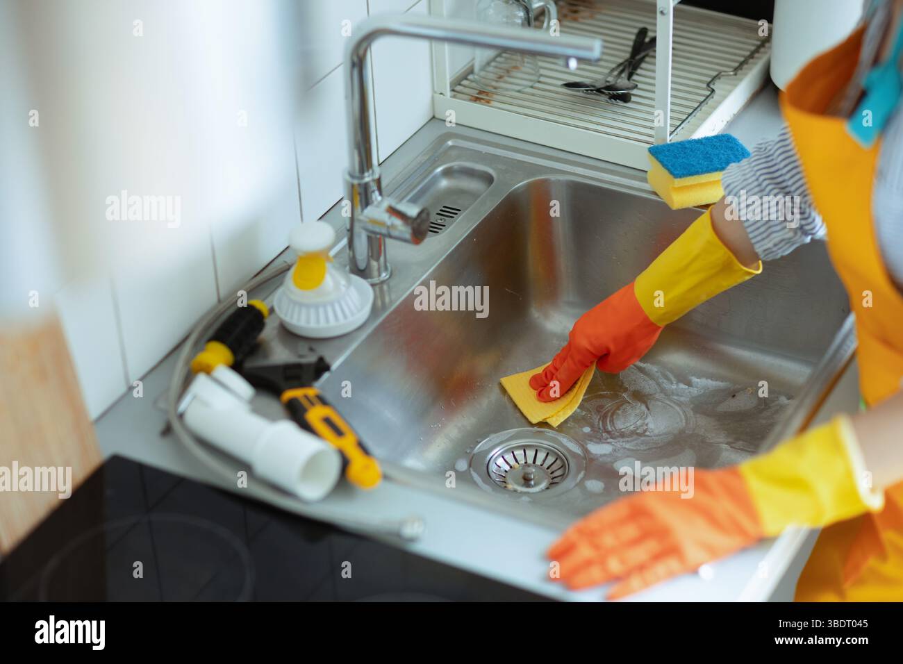 Gloved hands scour a sudsy stainless sink, plumbing tools nearby, showing ongoing effort to clean and unblock a stubborn kitchen drain. Stock Photo