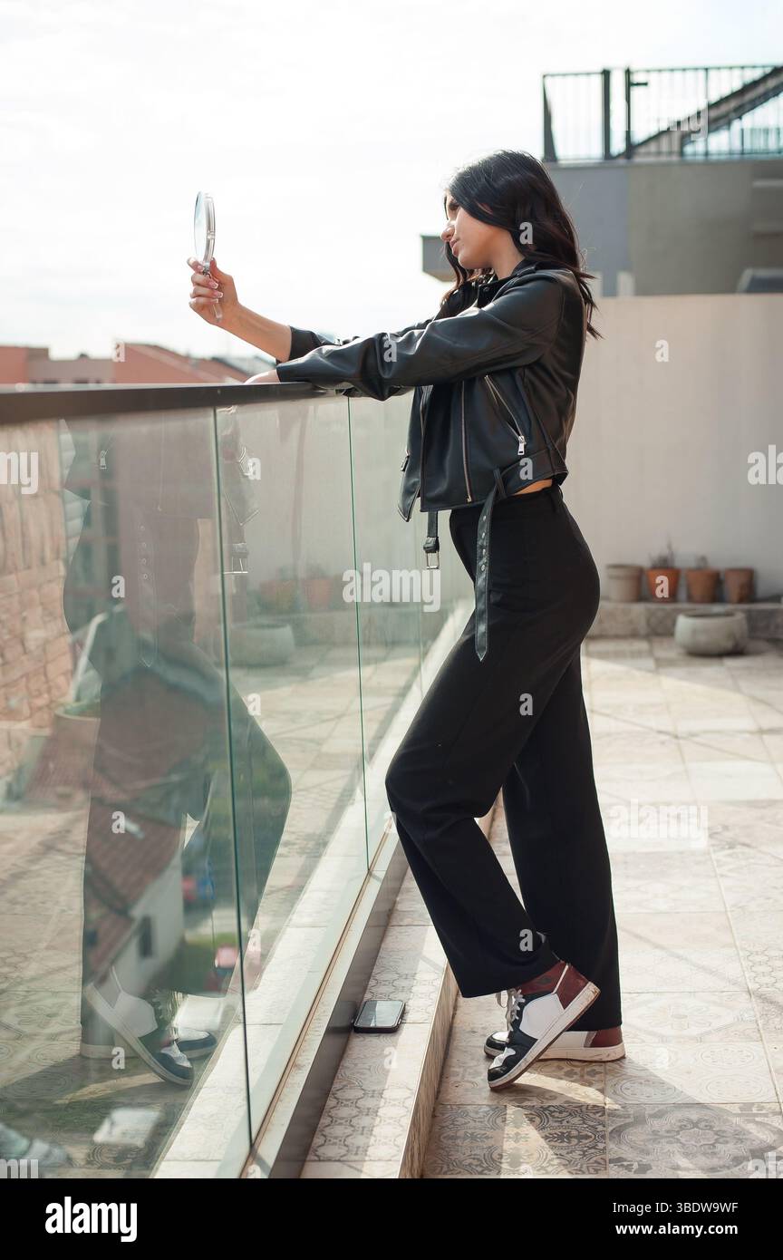A young, fashionable woman is posing near a glass barrier, looking at herself in a handheld mirror. She is on a rooftop during the daytime, wearing bl Stock Photo