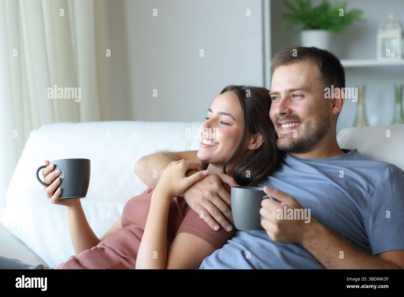 Happy couple drinking tea looking away sitting on a couch in a house interior Stock Photo