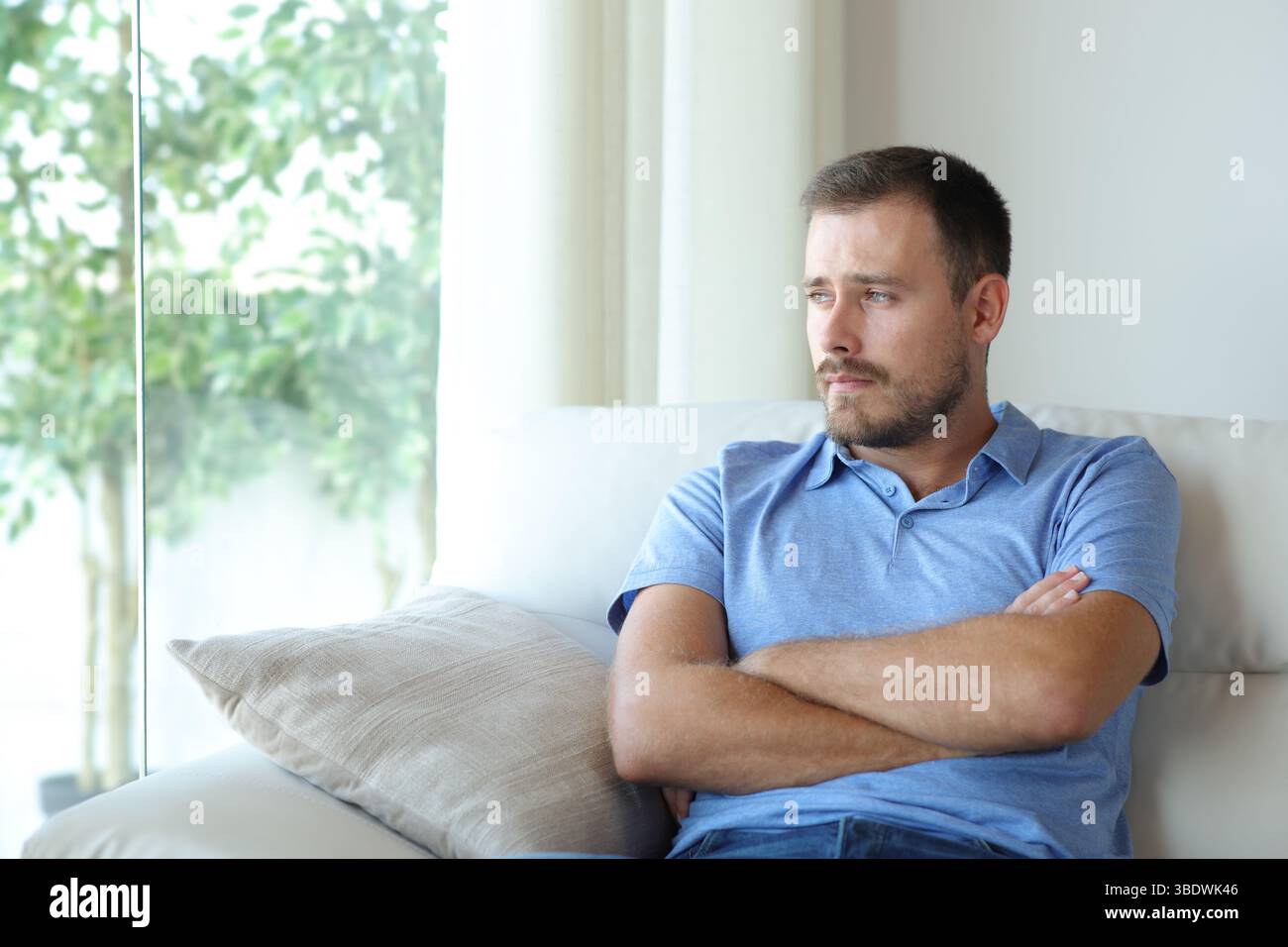 Pensive sad man looking through a window sitting on a couch at home Stock Photo