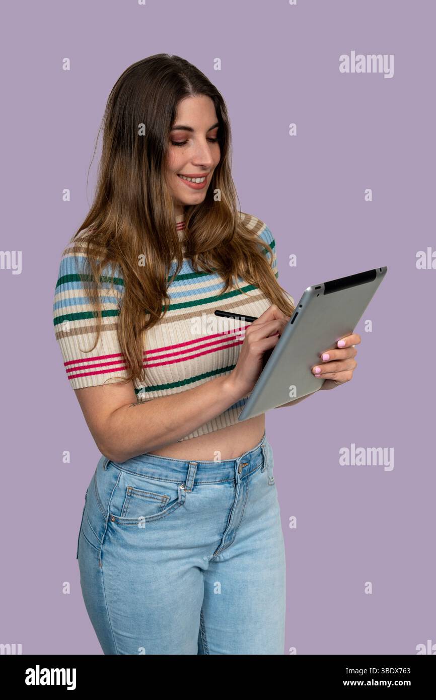 Smiling young woman using a tablet with stylus pen, working or studying on a purple background Stock Photo
