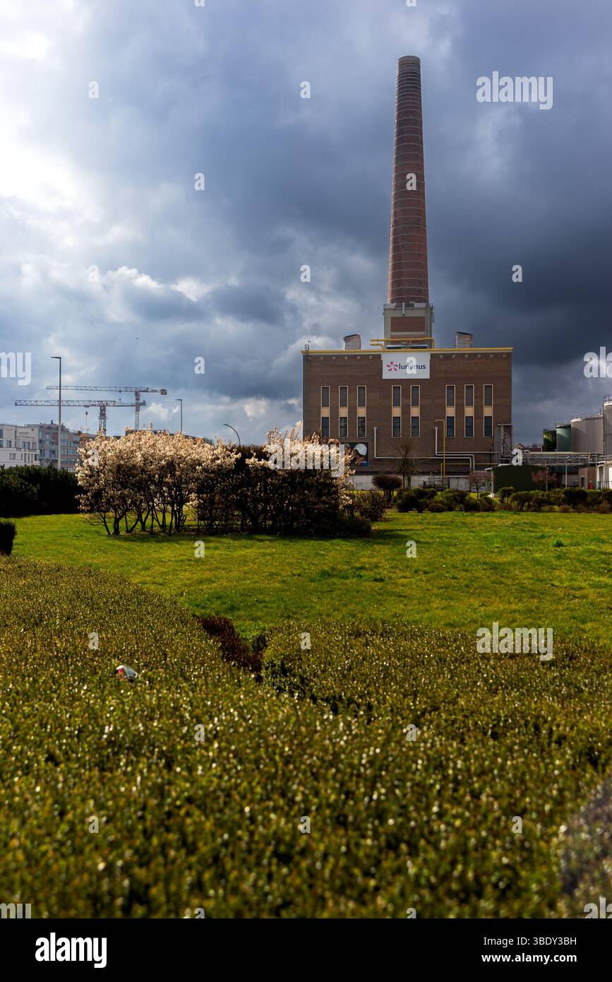 30.03.2025 Gent, Belgium, a tall brick smokestack rises from a Luminus power station under dramatic cloudy skies with green lawns, trimmed bushes, and Stock Photo