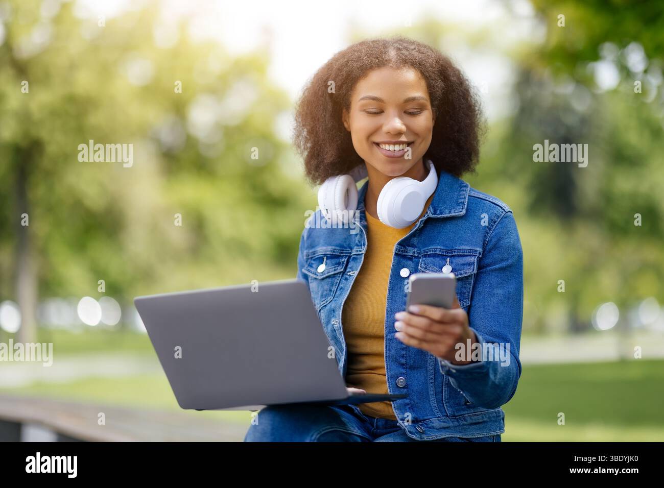 Happy Black student woman using smartphone and laptop outdoors Stock Photo