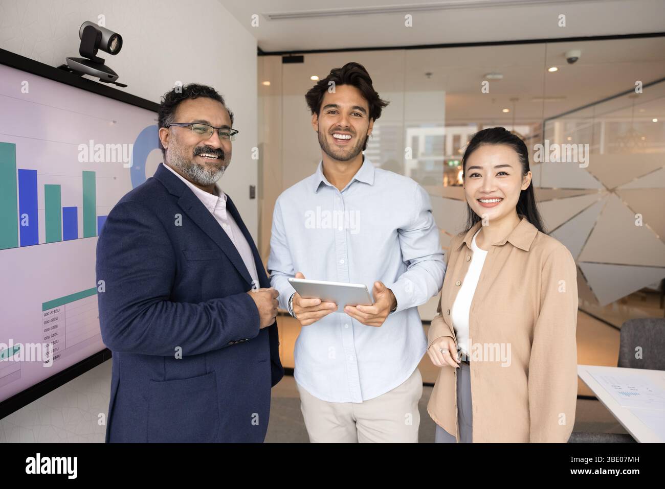 Three professionals standing in office next to screen displaying graphs Stock Photo